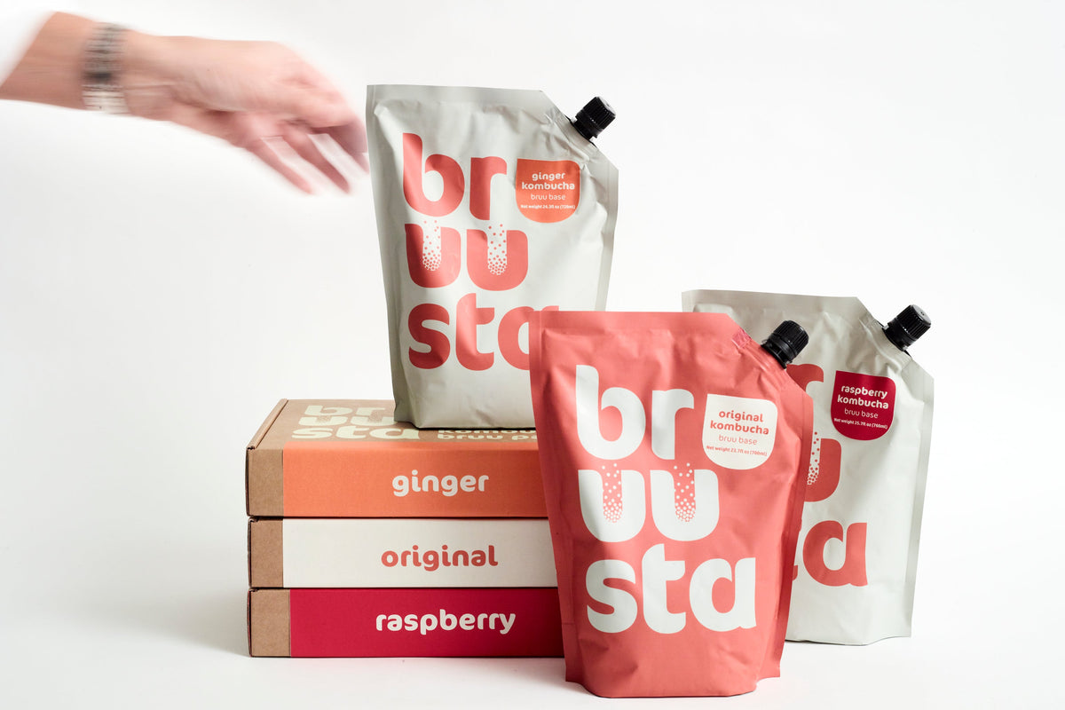 Pouches of bruu base builders in various flavors including ginger, original, and raspberry placed on a box with a person's hand reaching for one of the pouches.