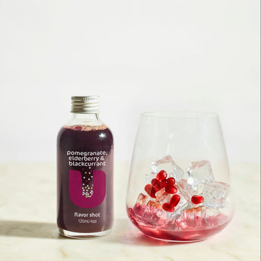 Bottle of pomegranate blackcurrant flavor shot next to a glass with pomegranate seeds and ice on a light background