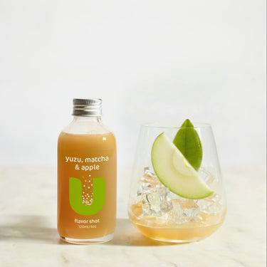 Bottle of yuzu, matcha & apple flavor shot next to a glass with ice and a slice of apple on a light background