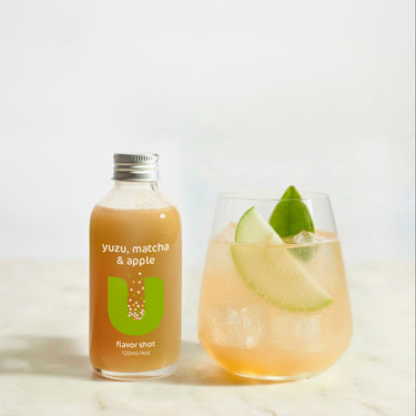 Bottle of yuzu, matcha & apple flavor shot next to a glass of the drink with apple wedges on a white background