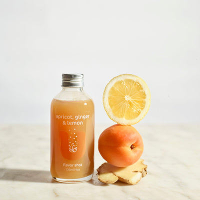 Bottle of apricot, ginger, and lemon flavored shot with a lemon, apricot, and ginger root on a light background