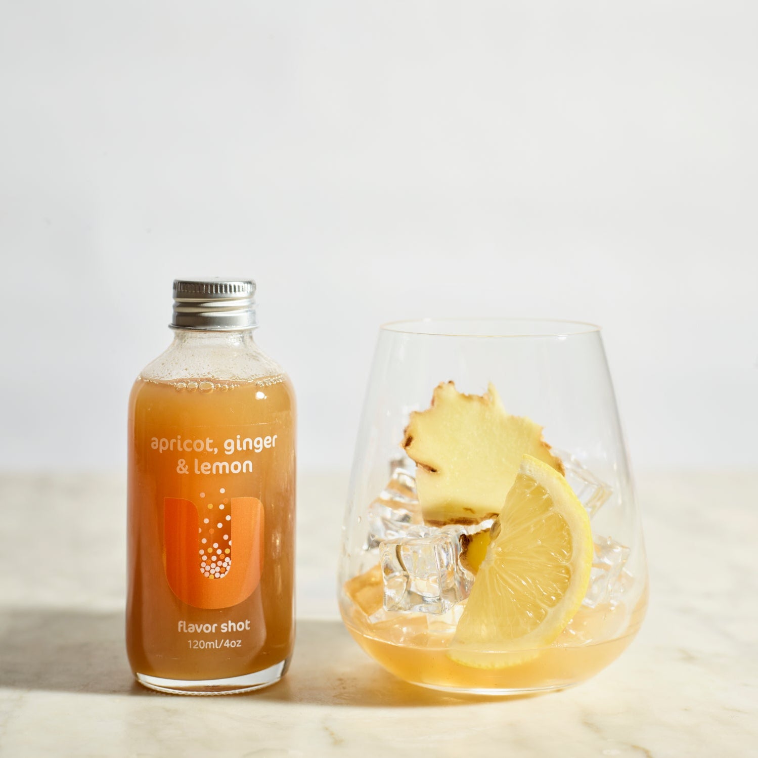 Bottle of 'Apricot, Ginger & Lemon' flavor shot next to a glass with a slice of lemon and a piece of ginger on a light background.