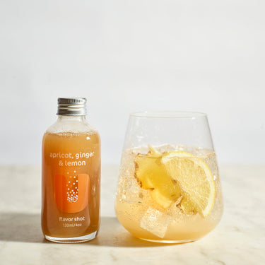 Bottle of 'Apricot, Ginger & Lemon' flavor shot next to a glass with lemon slices and ice on a light background