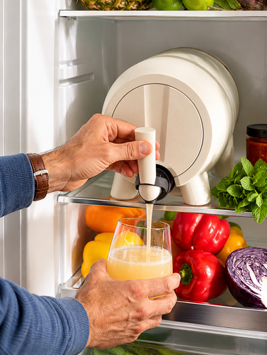 Person pouring kombucha from a white keg into a glass in front of an open refrigerator filled with fruits and vegetables.
