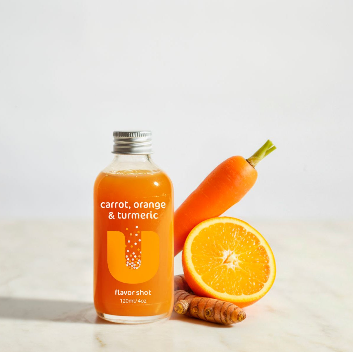 Bottle of carrot, orange, and turmeric flavorshot with a carrot, orange, and turmeric root on a light background