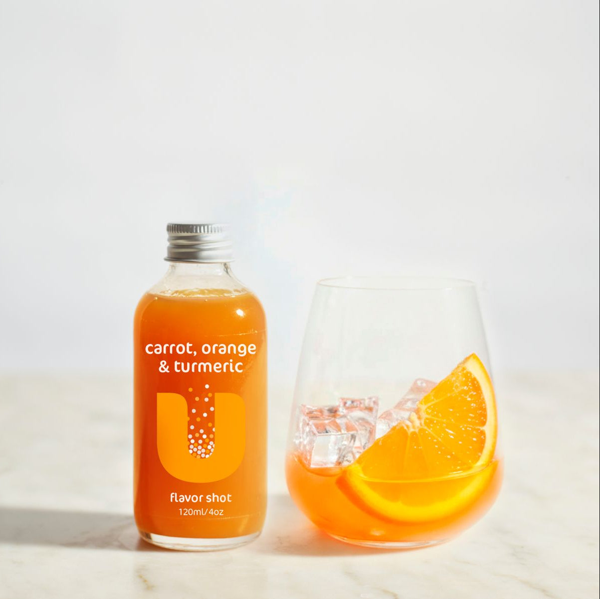 Bottle of 'carrot, orange & turmeric' flavorshot next to a glass with orange slices on a light background