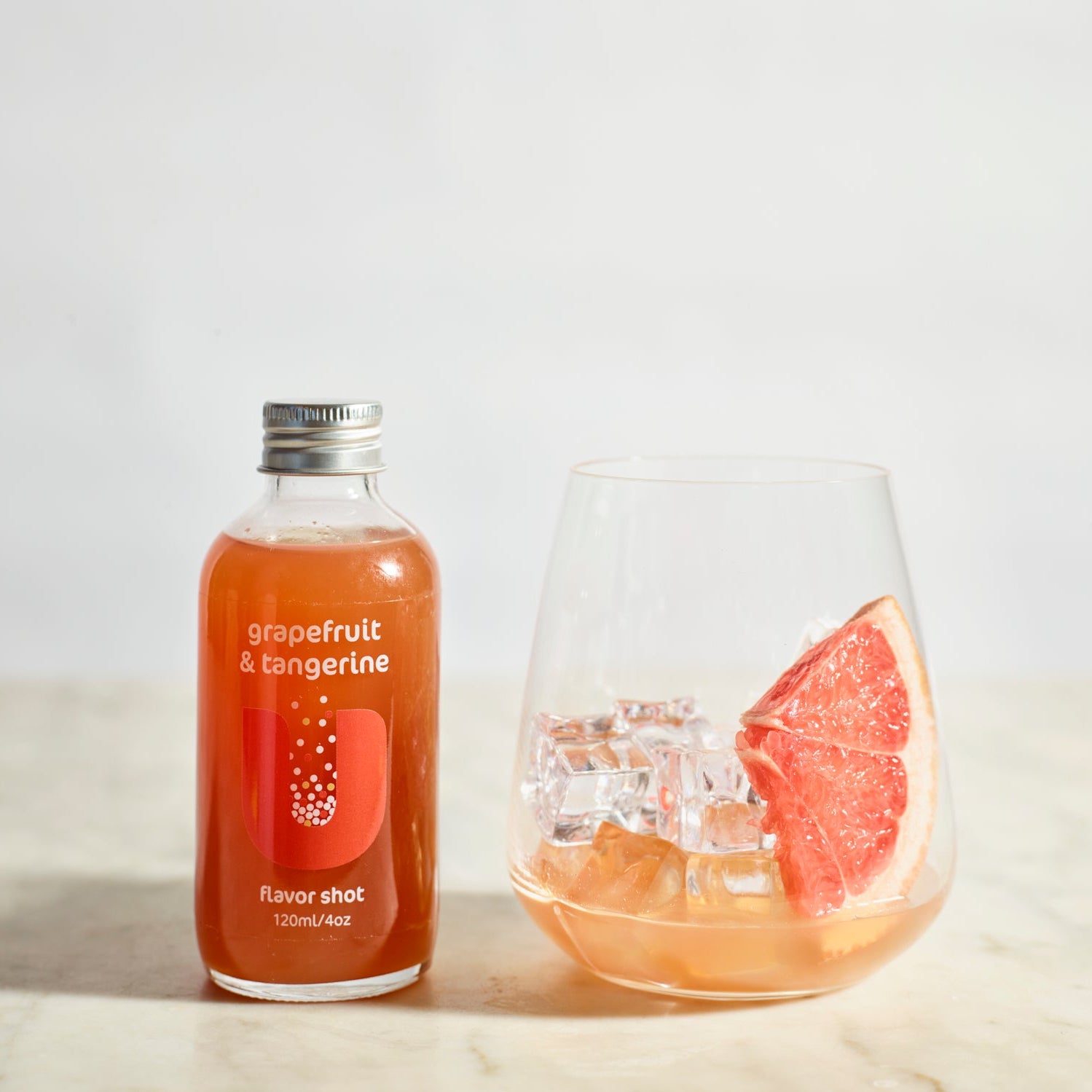Bottle of grapefruit and tangerine flavor shot next to a glass with a drink and grapefruit slice on a light background
