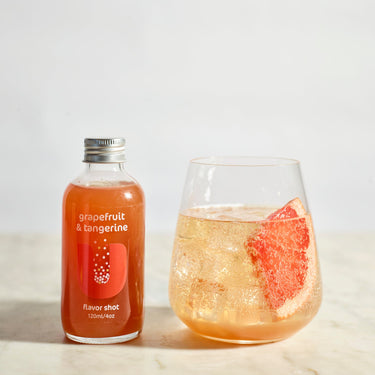 Bottle of grapefruit and tangerine flavor shot next to a glass of the drink with a slice of grapefruit on a light background