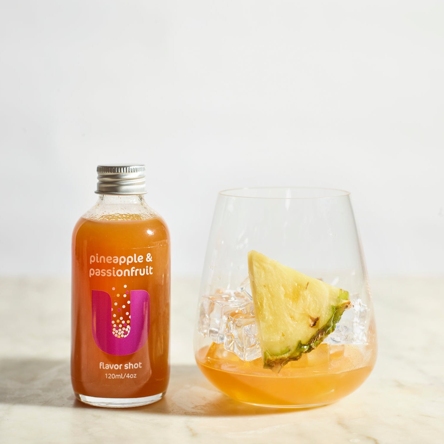 Bottle of pineapple & passionfruit juice shot next to a glass with a slice of pineapple on a white background