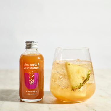 Bottle of pineapple & passionfruit flavor shot next to a glass of the drink on a light background