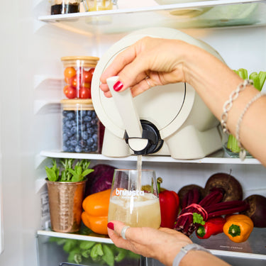 A person's hands pour a light-colored beverage from a kombucha home brewing and on tap device into a glass inside an open refrigerator shelf filled with fresh vegetables and jars.