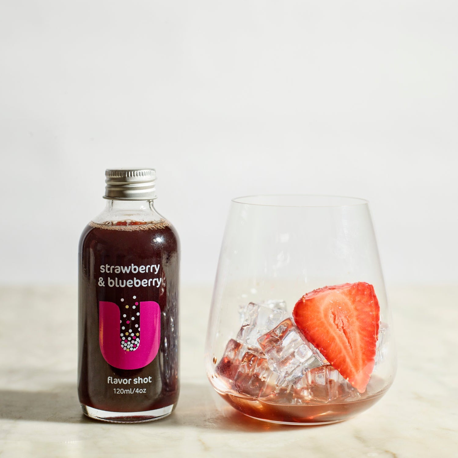 Bottle of strawberry & blueberry flavored shot next to a glass with ice and a strawberry on a light background