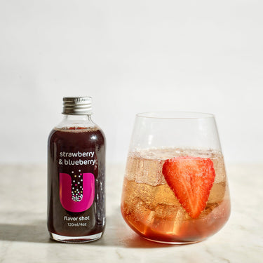 Bottle of strawberry & blueberry flavored shot next to a glass of the same flavor on a light background